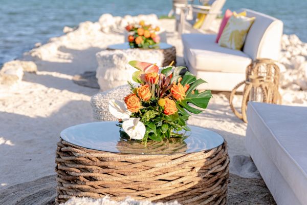 A seaside lounge area with stylish seating, cushions, and decorative flowers on woven tables overlooks a calm ocean.