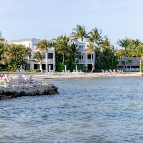 A scenic view of a seaside resort features beachfront buildings, palm trees, and lounge chairs, with calm water in the foreground ending the sentence.