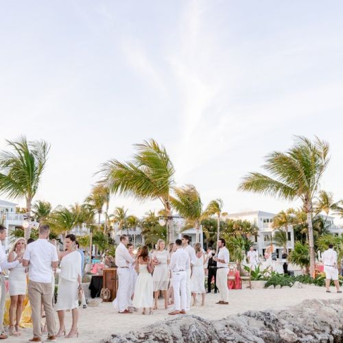 A group of people dressed in white are socializing at a beachside gathering with palm trees and buildings in the background.