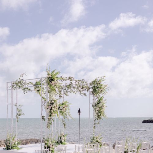 An outdoor wedding setup by the sea with a white, decorated arch and rows of white chairs, under a clear, blue sky with scattered clouds.