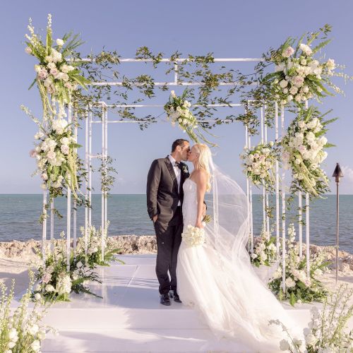 A couple kisses under a decorated wedding arch by the sea, surrounded by flowers and clear skies. The ceremony appears romantic and serene.