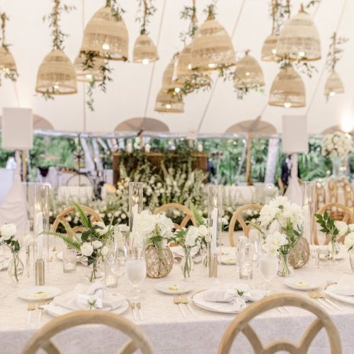 A beautifully decorated wedding reception with white floral arrangements, elegant table settings, and hanging lanterns under a large canopy tent.