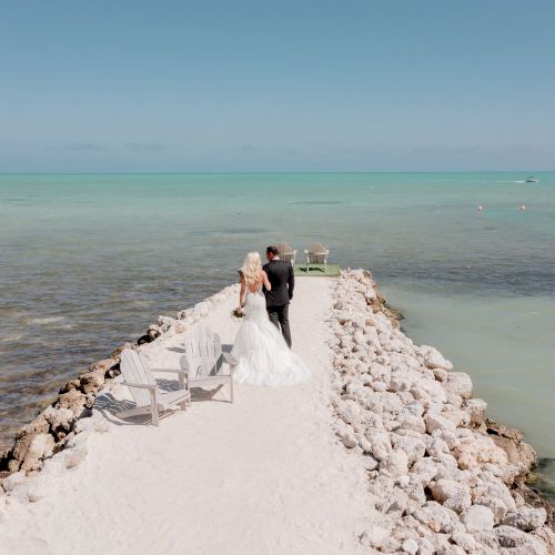 A couple in wedding attire stands on a rocky pier overlooking a tranquil sea, with two empty chairs facing the water.