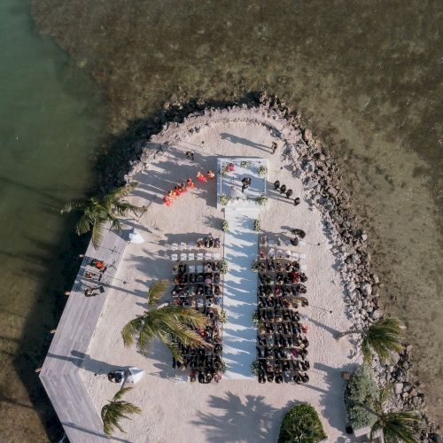 Aerial view of a small peninsula with a setup for an event, featuring rows of chairs, a pathway, and palm trees, surrounded by water and rocky edges.