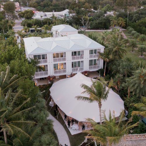 Aerial view of a large white building with a tent set up outside surrounded by lush greenery and palm trees, typical of a tropical setting.