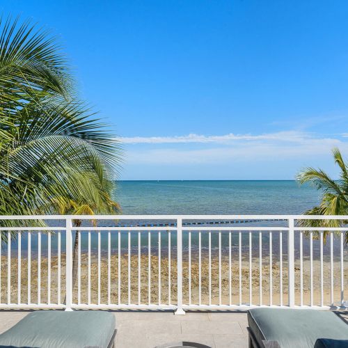 A sunny beach view with palm trees, a white railing, and lounge chairs on a balcony overlooking the calm sea and clear blue sky.