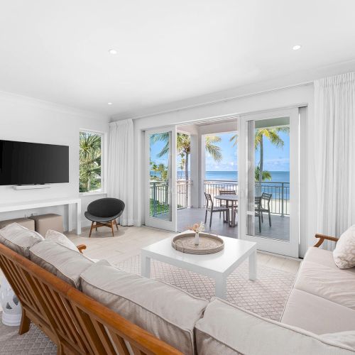 A bright living room with neutral furniture, a wall-mounted TV, and sliding doors opening to a balcony with an ocean view and palm trees.