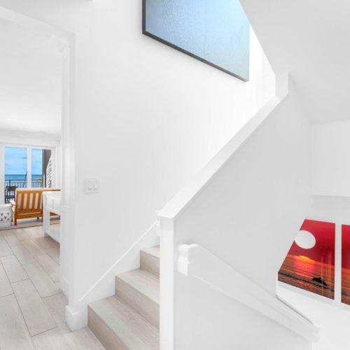 A bright, modern interior with a staircase, art on the walls, and a view of the ocean through large windows in the living area.