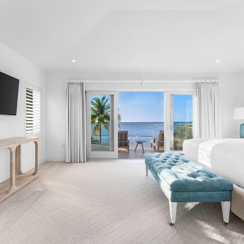 Bright bedroom with ocean view, featuring a bed, blue bench, TV, and large glass doors opening to a balcony with palm trees and blue skies.