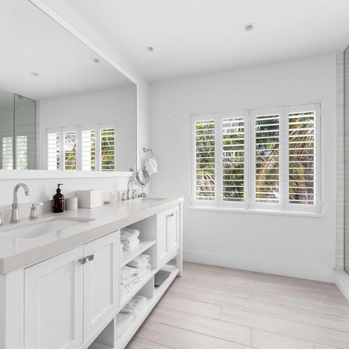 A modern bathroom with a double sink vanity, large mirror, glass shower, and white shutters on the window.