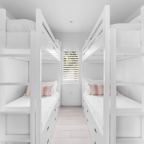 The image shows a bright, white room with two sets of bunk beds, parallel to each other, and a window with blinds.