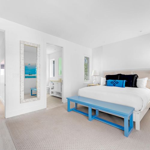 A bright bedroom with a white bed, blue bench, and mirrored wall. The space is airy and modern, featuring minimalist decor and natural light.