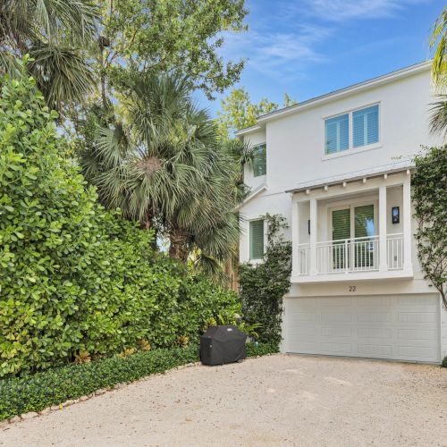 A two-story white house with a balcony and garage surrounded by lush greenery and palm trees under a clear blue sky.