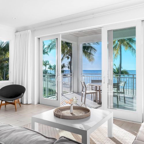 A modern living room with a balcony view of the ocean and palm trees, featuring sleek furniture and large sliding glass doors.