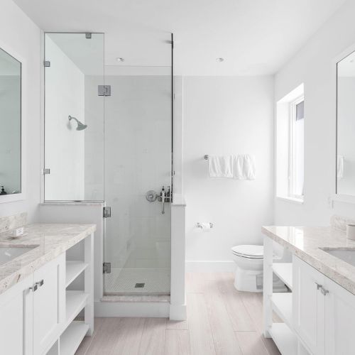 The image shows a modern white bathroom with double sinks, large mirrors, a glass shower, and a toilet. It has a clean and minimalist design.