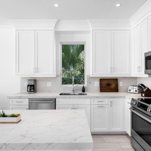 A modern kitchen with white cabinets, stainless steel appliances, and a marble island. Two windows show lush greenery outside.