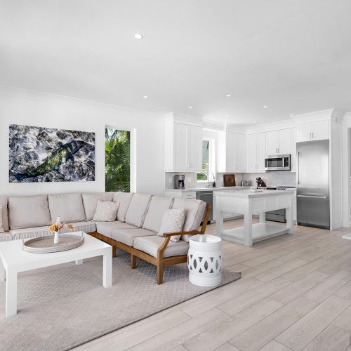 A modern, bright living room with a large sofa, coffee table, and an open kitchen dining area, featuring white and light wood decor.