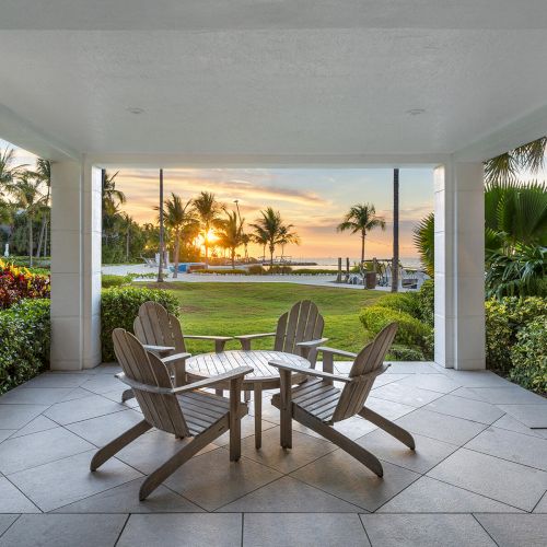 A patio with four wooden chairs around a table, overlooking a grassy area with palm trees and a sunset view over the ocean.