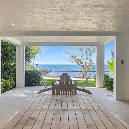 A covered patio area opens to a view of the ocean, framed by greenery, with two wooden chairs facing the water under a bright sky.