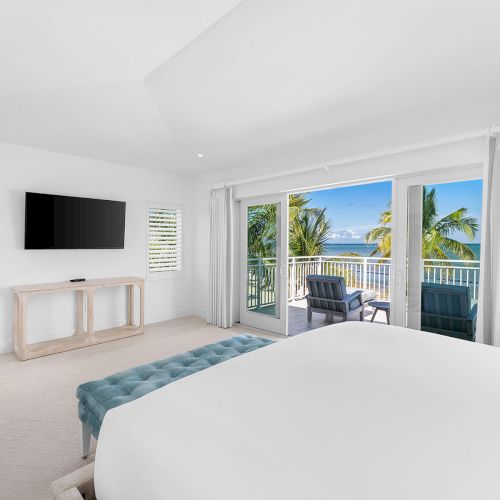 A bright bedroom with a large bed, TV, and a balcony view of palm trees and the ocean, featuring white and blue decor elements.