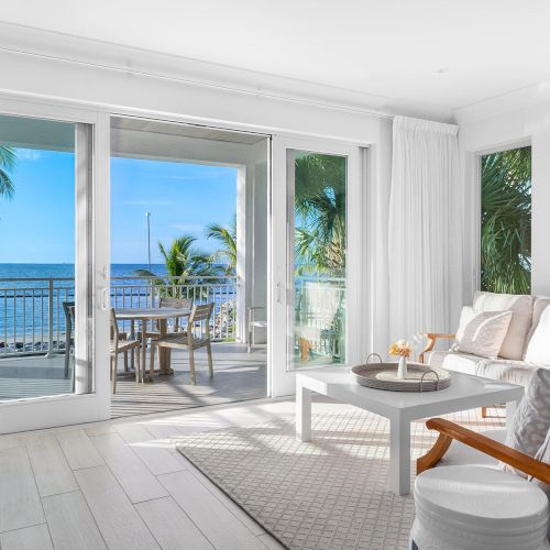 A bright, modern living room with white decor opens to a balcony overlooking the ocean and palm trees.