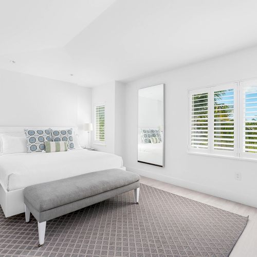 A bright, modern bedroom with a large bed, bench, nightstands, and a full-length mirror, features white walls and a window with shutters.