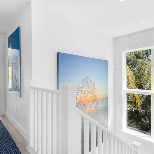 A bright hallway with white walls, staircase, ocean-themed artwork, a window showcasing palm trees, and a glimpse into a coastal-themed room.