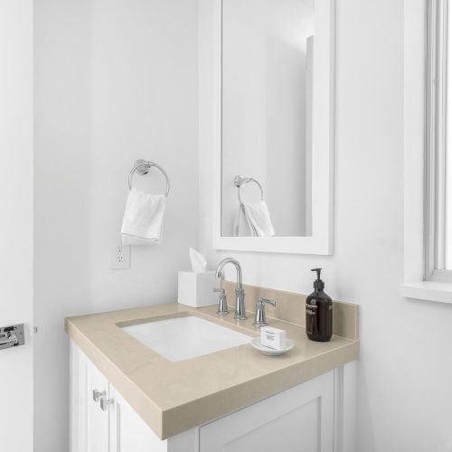 A small, minimalist bathroom with a white countertop, sink, faucet, mirror, soap dispenser, towel, tissues, and a window beside the mirror.