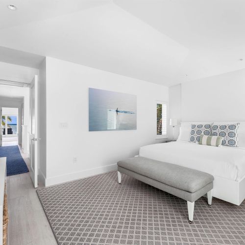 A modern bedroom with a white bed, gray bench, patterned rug, and TV. There's wall art and a hallway leading to another room.