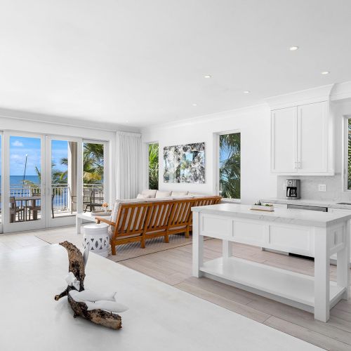 A bright, modern living space with a kitchen island, white cabinetry, wooden furniture, and a view of the ocean through large glass doors.