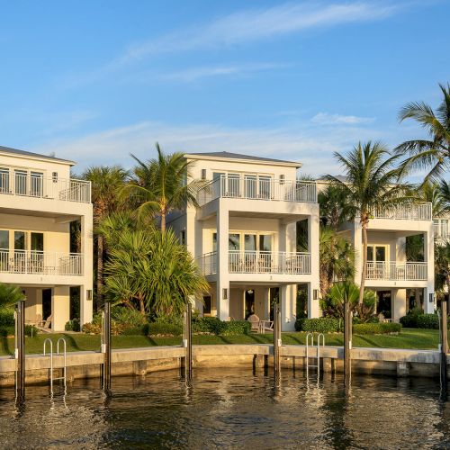 The image shows a row of modern waterfront homes surrounded by palm trees, with a calm waterway in the foreground, under a clear sky.