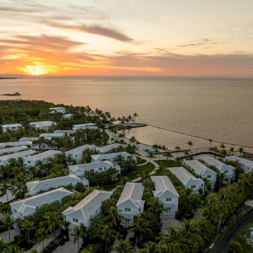 A coastal sunset view with white-roofed buildings surrounded by greenery, near a calm body of water.