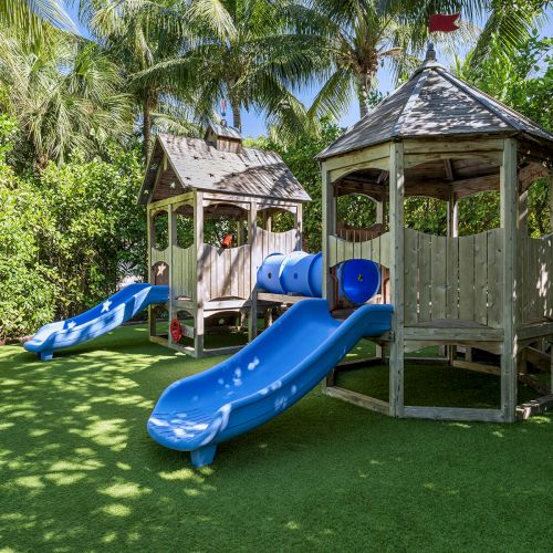 A playground with two wooden structures, blue slides, and a tunnel, surrounded by green trees and grass, under a bright blue sky.