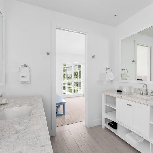 A modern bathroom with dual sinks, marble countertops, large mirrors, and an adjoining room with a blue bench and window shutters.