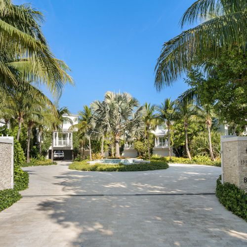 Luxurious gated entrance with palm trees and a fountain, leading to elegant white buildings in a tropical setting.