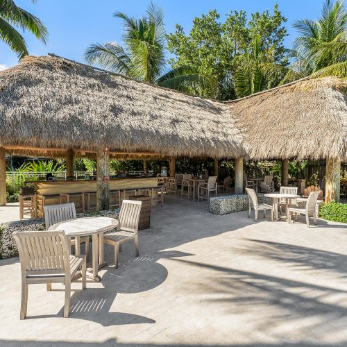 An outdoor seating area with wooden tables and chairs under a thatched roof, surrounded by tropical trees and a clear blue sky.