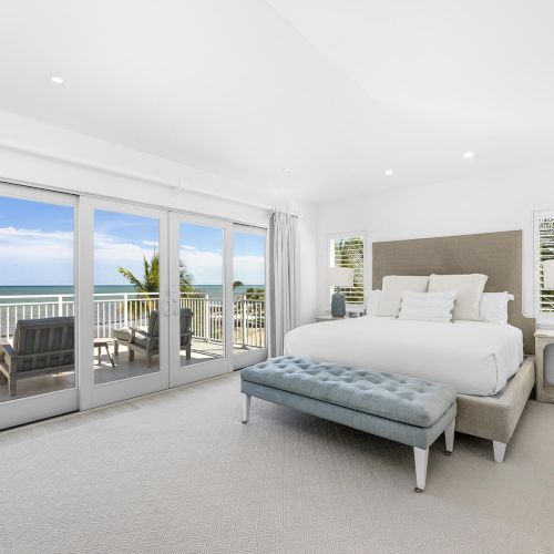 This image shows a bright bedroom with a large bed, elegant decor, and a view of the ocean through glass doors leading to a balcony.