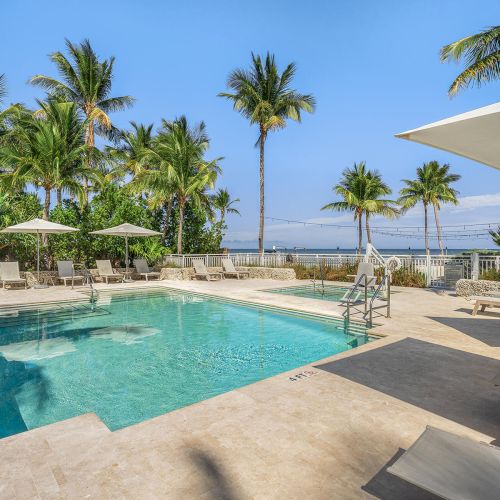 A tranquil outdoor swimming pool area with lounge chairs, umbrellas, and palm trees, offering a view of the ocean and a clear blue sky.