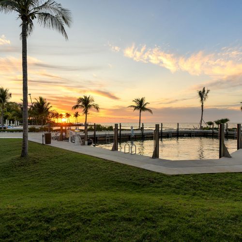 A serene waterfront view featuring palm trees, a small harbor, and a vibrant sunset over calm waters, with well-maintained grassy areas.