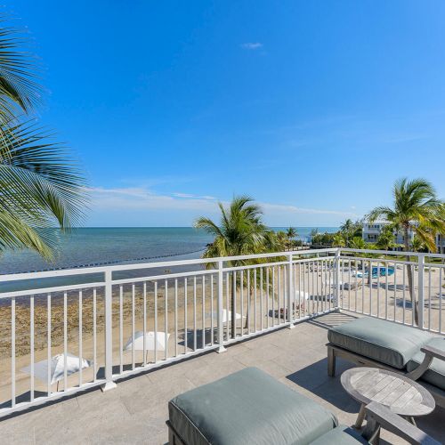 A beachfront view with palm trees, lounge chairs, and a clear blue sky on a sunny day, seen from a balcony.