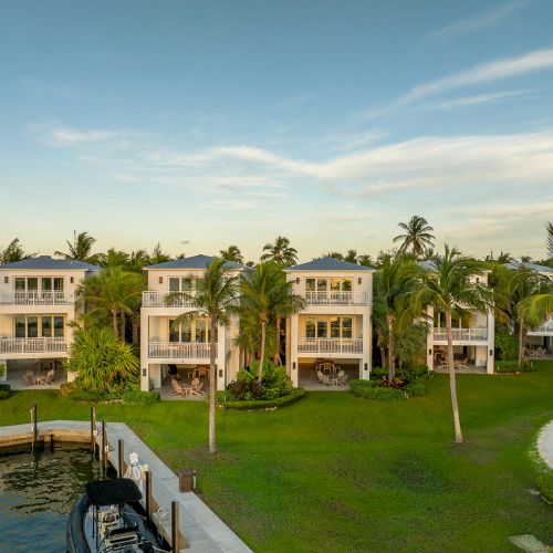 The image shows a series of white houses surrounded by palm trees, set beside a waterway with docks, under a blue sky with clouds.