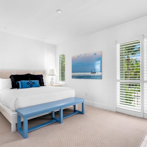 A bright bedroom with a large bed, blue bench, white walls, artwork, and windows with blinds allowing sunlight in.