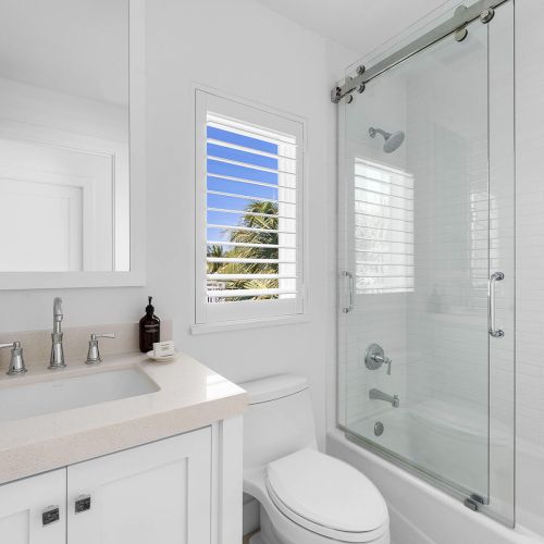 A bright, modern bathroom with a sink, mirror, toilet, and glass shower. A window offers a view of clear skies and palm trees.