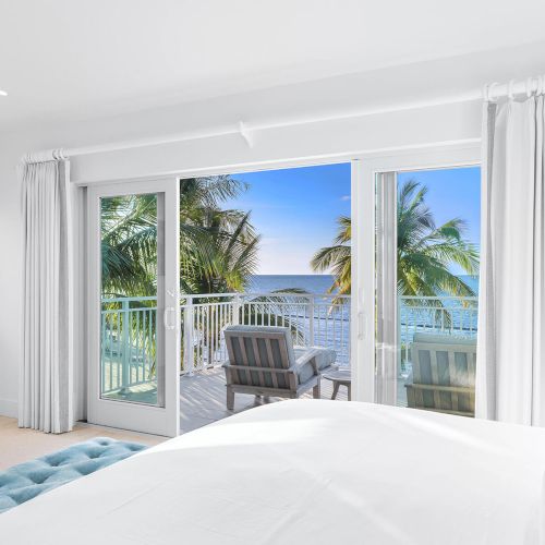 A serene bedroom with white decor opens to a balcony view, featuring palm trees and a tranquil ocean scene beyond glass doors.