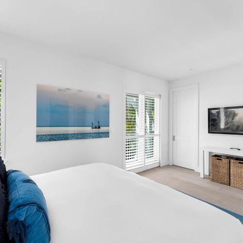 A modern bedroom with a large bed, coastal artwork, TV, wicker storage, and natural light from windows with white shutters.