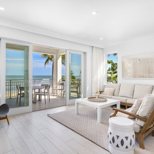 A bright living room with a white couch, coffee table, and a chair opens to a balcony with ocean views through sliding glass doors.