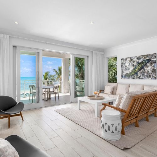 A bright, modern living room with ocean view through glass doors, wooden furniture, and wall art. Tropical setting with sea and palm trees outside.