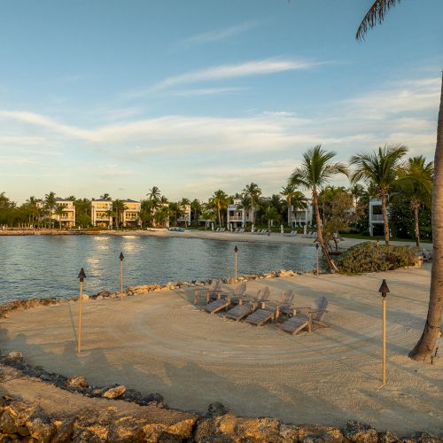 A serene beach scene with lounge chairs, tiki torches, palm trees, and calm waterfront, under a clear sky.