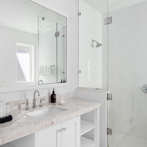 A modern bathroom features a glass shower, marble countertop with sink, large mirror, and minimalistic decor with a soap dispenser on the counter.