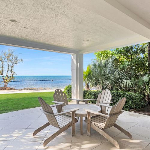 A covered patio with four wooden chairs and a small table overlooks a grassy area and a beach with a calm sea view in the background.